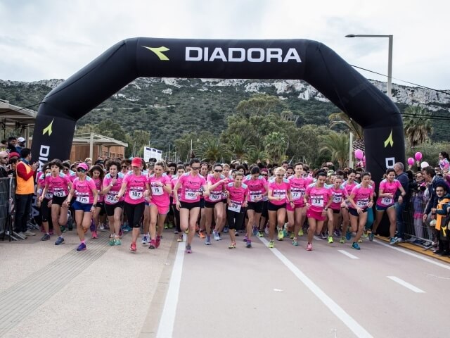 Gruppo di corridori in magliette rosa sotto un arco "DIADORA", con spettatori e paesaggio di alberi e colline sullo sfondo.