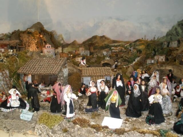 Diorama di un villaggio rurale con persone in abiti tradizionali, case in pietra e legno, e un paesaggio montano sullo sfondo.