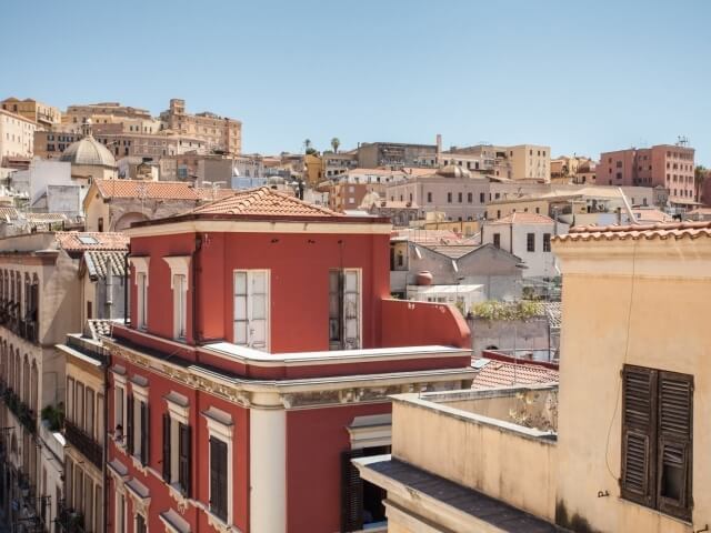 Un vivace paesaggio urbano con un edificio rosso al centro, circondato da edifici di varie dimensioni e un cielo blu.