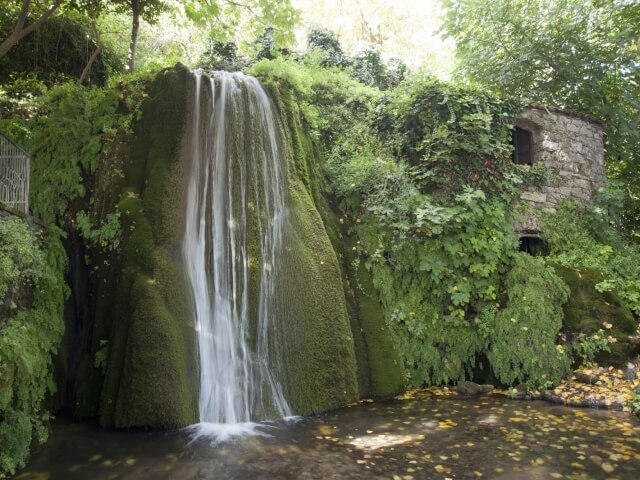 Cascata serena su rocce coperte di muschio, con una piccola struttura in pietra e vegetazione lussureggiante circostante.