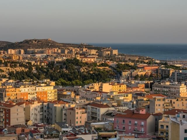 Vista di una città collinare con edifici a tetto rosso, alberi verdi e un grande corpo d'acqua sullo sfondo sotto un cielo sereno.