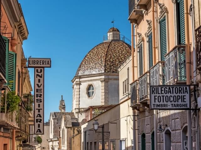 Scena urbana serena con edifici, un edificio centrale con cupola, insegne e cielo blu senza nuvole.