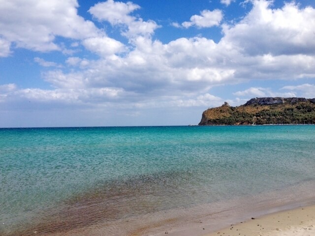 Spiaggia serena con acqua tranquilla, sabbia bagnata, collina verde in lontananza e cielo blu punteggiato di nuvole bianche.