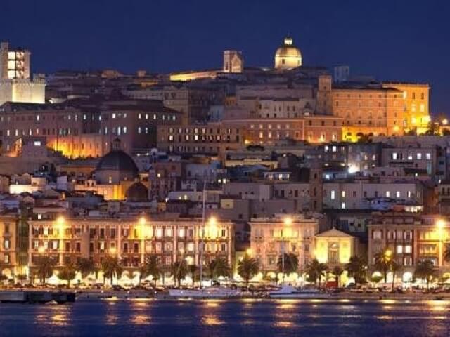 Paesaggio urbano notturno con acqua in primo piano, edifici illuminati e palme lungo la riva, atmosfera tranquilla.