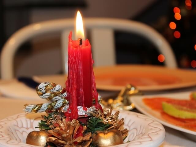 Tavola di Natale con candela rossa, piatto bianco decorato, pigna, ornamenti dorati e albero di Natale sullo sfondo.