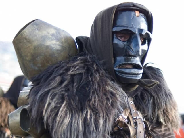 Persona con maschera nera e cappuccio di pelliccia, zaino cilindrico metallico, sfondo sfocato di cielo bianco.