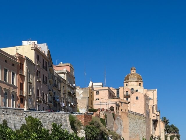 Una fila di edifici chiari su un molo di pietra, con un grande edificio a cupola e vegetazione lussureggiante.