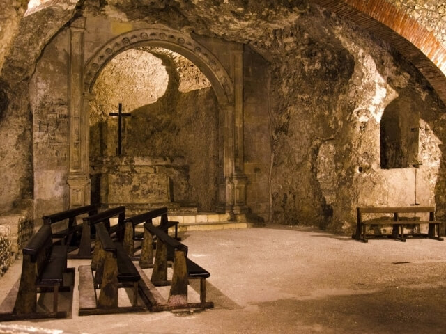 Interno di una cappella storica con panche in legno, archi in pietra e un crocifisso nero su una piattaforma.