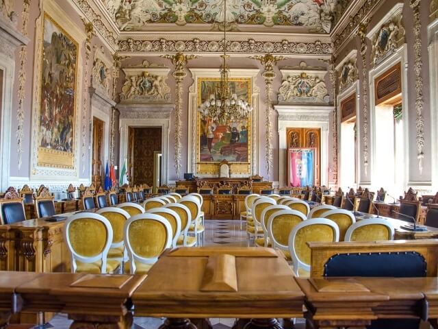 Una grande sala ornata con tavoli e sedie, soffitto decorato e dipinti, suggerendo un ambiente formale e prestigioso.