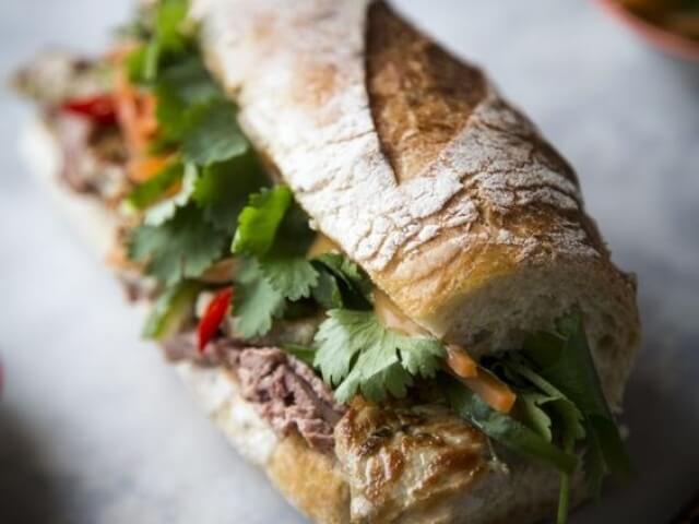 Un primo piano di un panino baguette con tonno, carote e peperoni, su un tavolo grigio sfocato sullo sfondo.