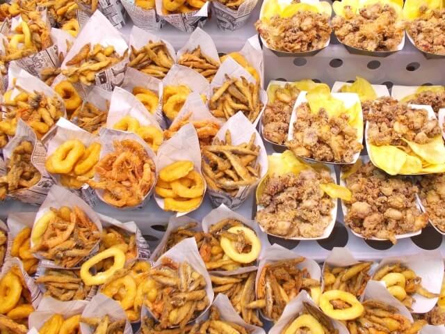 Immagine di cibo fritto, tra cui patatine e anelli di cipolla, serviti in contenitori di carta su un tavolo.