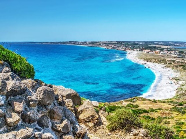 Spiaggia serena con acqua blu, sabbia bianca, rocce grigio-marroni e persone che nuotano e prendono il sole.