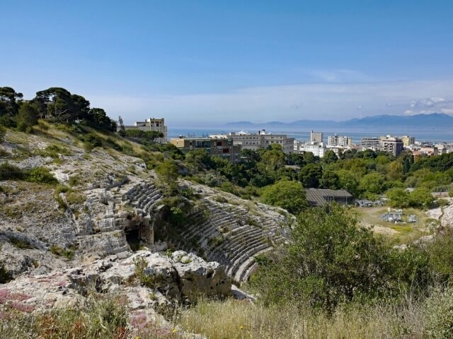 Vista aerea di un teatro scolpito nella collina, circondato da una città costiera e montagne, in una giornata serena.