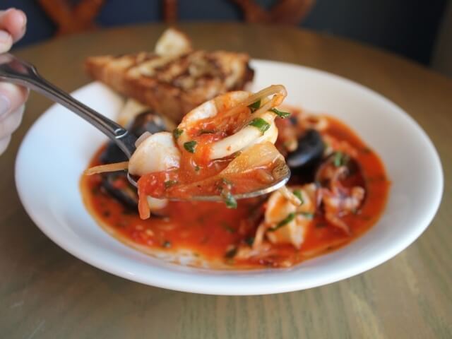 Un piatto bianco di frutti di mare in salsa rossa, con pane tostato, su un tavolo rotondo, mentre una mano tiene un cucchiaio.