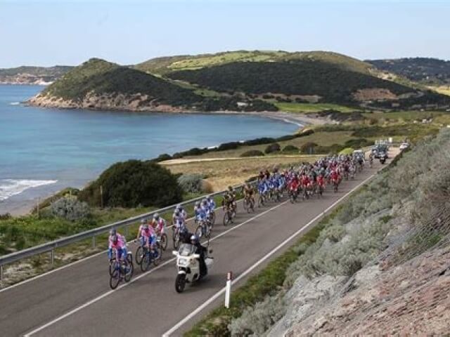 Un gruppo di ciclisti in abbigliamento colorato pedala lungo una strada costiera, con acqua a sinistra e colline sullo sfondo.