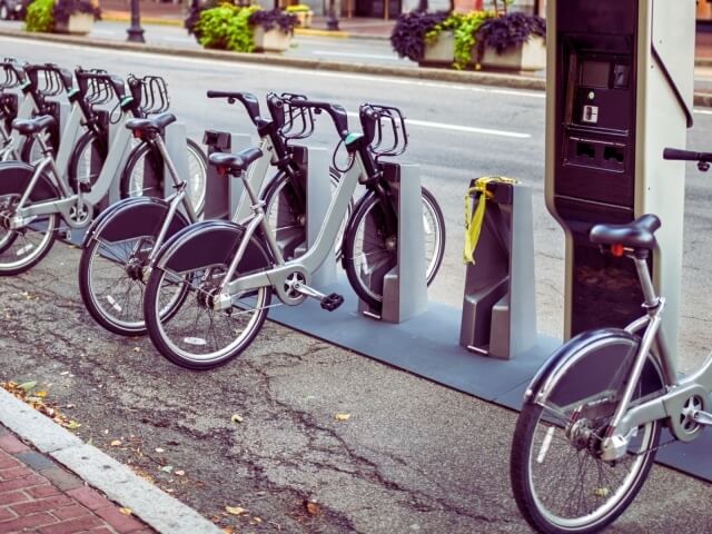 Una fila di biciclette bianche parcheggiate a una stazione, con un marciapiede e una strada asfaltata sullo sfondo.