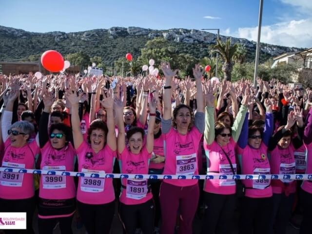 Un gruppo di donne in magliette rosa alza le mani, festeggiando in un evento con montagne sullo sfondo e cielo blu.