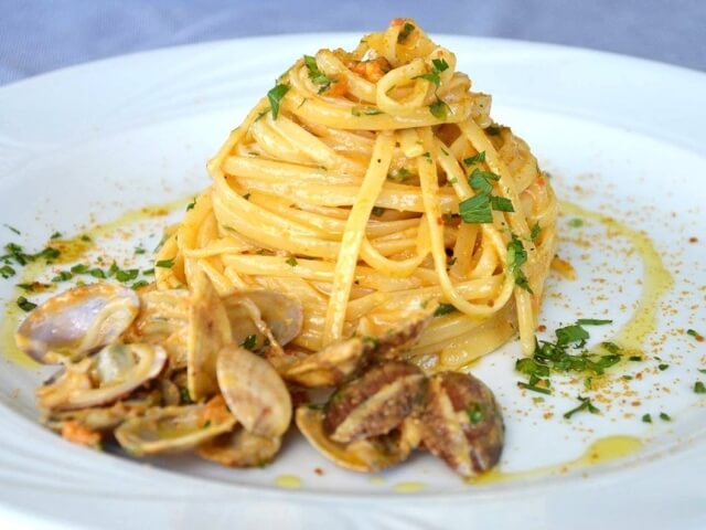 Un primo piano di pasta a forma di piramide con salsa gialla e prezzemolo, circondata da conchiglie vuote su un piatto.