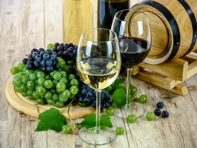 Disposizione di bicchieri di vino, una bottiglia, un barile di legno e uva su un tavolo di legno chiaro.