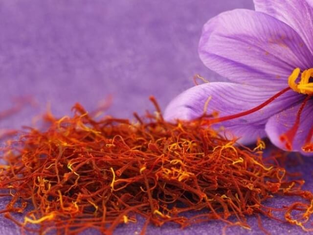 Primo piano di fili di zafferano arancioni e gialli con un fiore viola sullo sfondo.