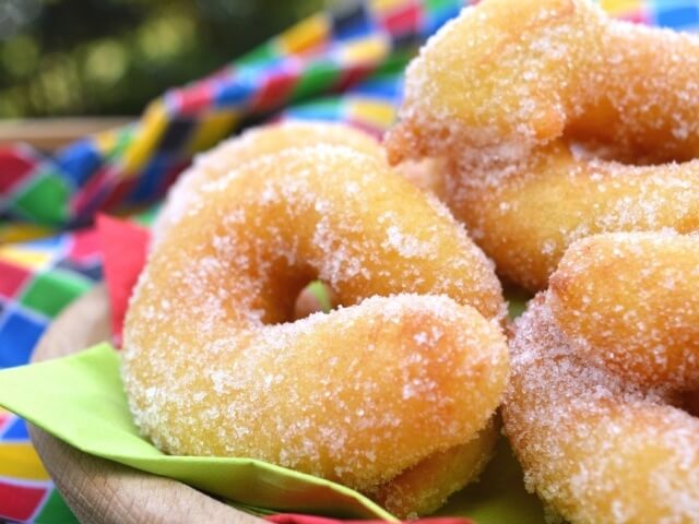 Cinque ciambelle zuccherate su un vassoio di legno, circondate da tovaglioli colorati, in un'atmosfera festiva.
