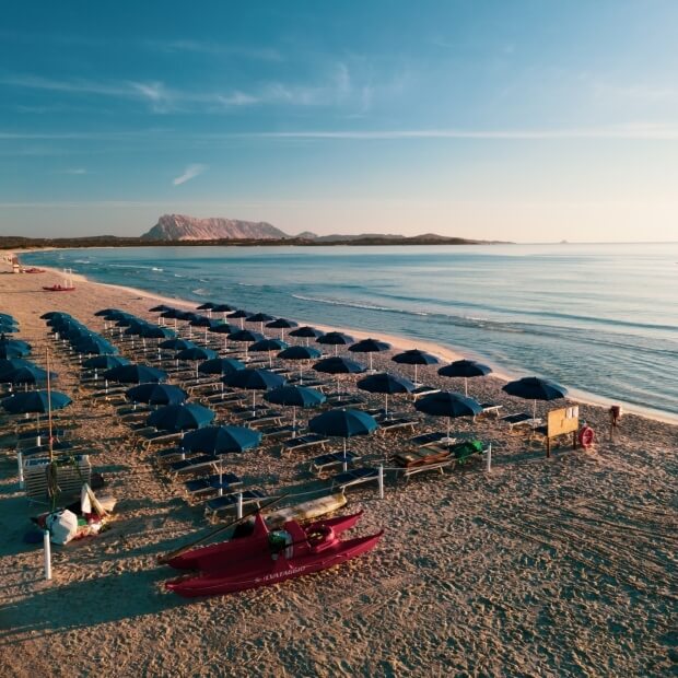 Spiaggia serena con sabbia, ombrelloni blu, kayak rossi e un'isola sullo sfondo sotto un cielo azzurro.