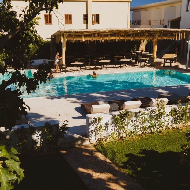 Area piscina serena con sdraio, ombrelloni, bar all'aperto e una persona che nuota, circondata da verde lussureggiante.