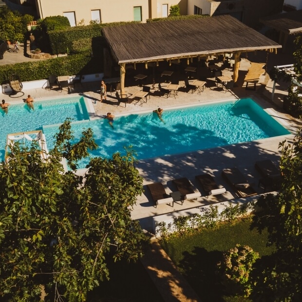 Piscina soleggiata circondata da vegetazione lussureggiante e persone sfocate, con struttura in legno e atmosfera rilassata.