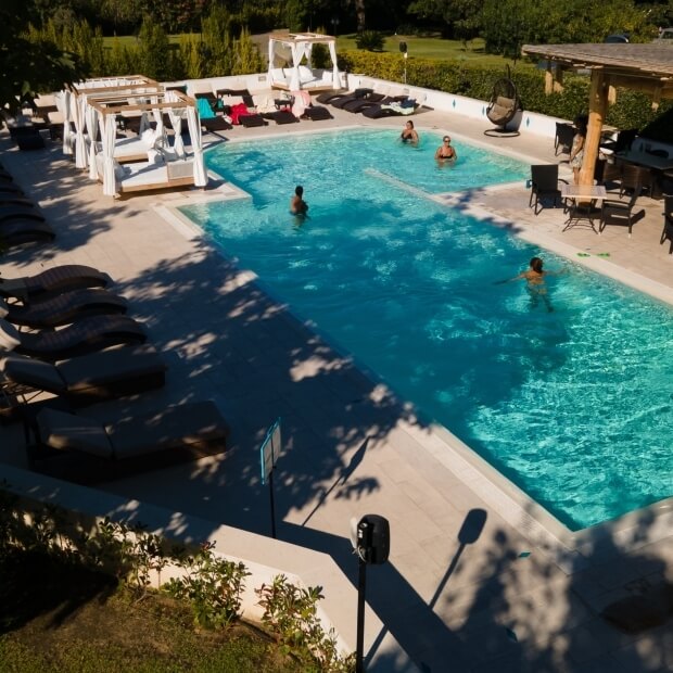 Una piscina serena con acqua blu, quattro persone, lettini e cabane, circondata da verde lussureggiante.