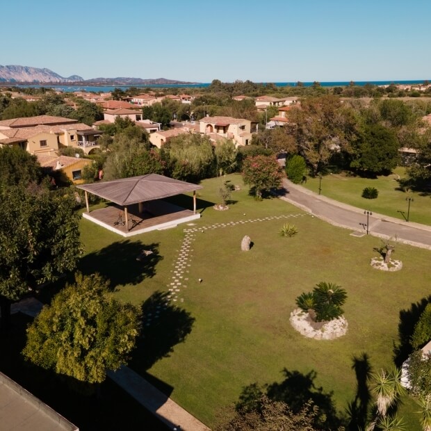 Vista aerea di un parco curato con prato, gazebo, alberi e un corpo d'acqua sullo sfondo, sotto un cielo sereno.