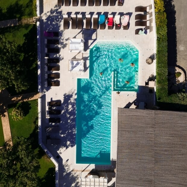 Vista aerea di una piscina rettangolare con sedie a sdraio, circondata da alberi e un edificio con tetto grigio.