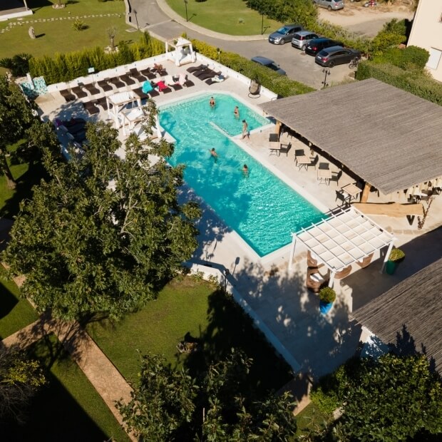 Vista aerea di una piscina rettangolare circondata da lettini e tavoli, con una struttura a tetto di paglia e un parcheggio.