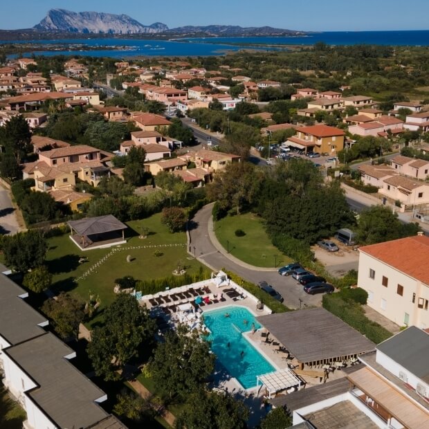 Vista aerea di un quartiere costiero con un grande hotel, piscina, alberi, case, montagne e un vasto corpo d'acqua.