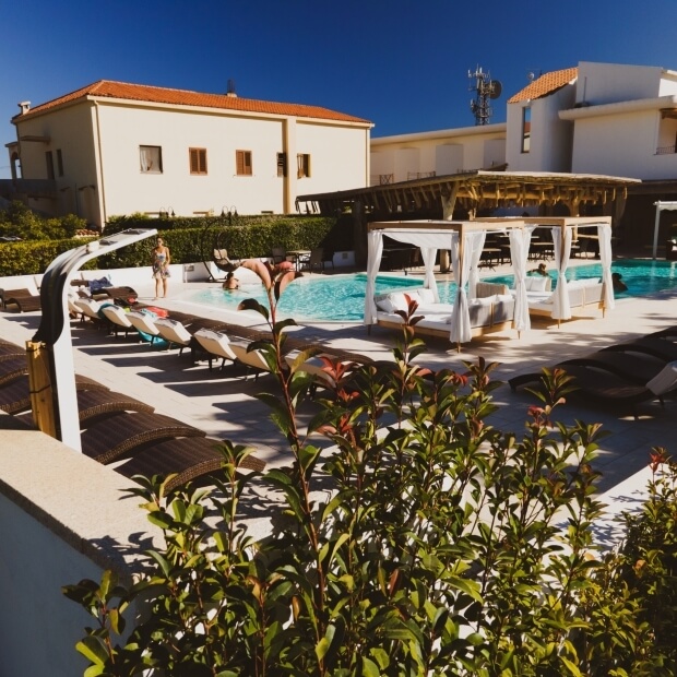 Area piscina serena con persone su lettini, gazebo a destra e edifici con vegetazione lussureggiante sullo sfondo.