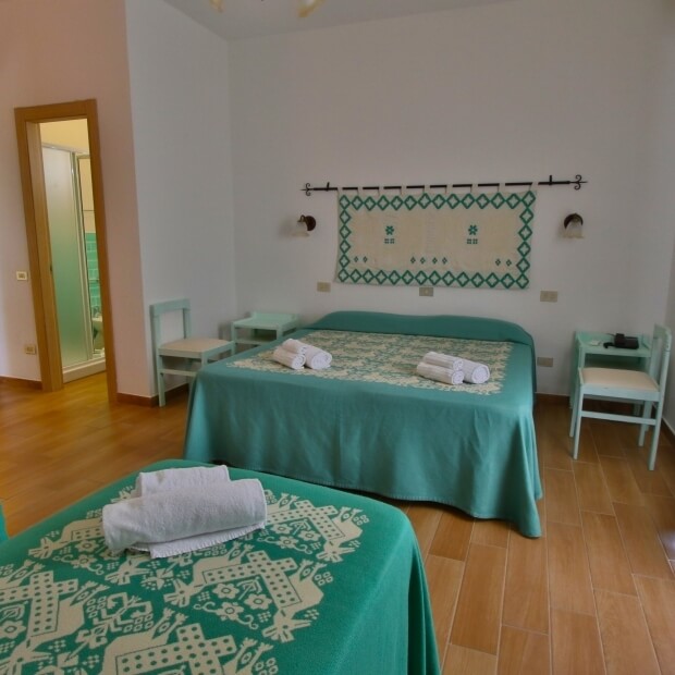 Camera accogliente con letti bianchi, coperte turchesi, arazzo decorativo e accesso a un bagno.