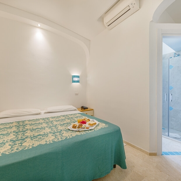 Camera serena con letto grande, coperta teal, due cuscini bianchi, e porta che conduce a un bagno con doccia.