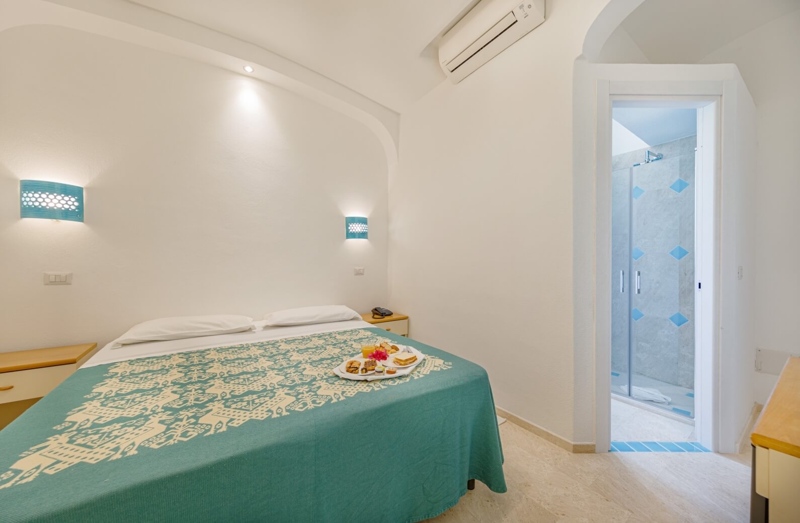 Camera d'albergo serena con letto, comodini, e bagno en-suite, decorata con coperte teal e dolci su un vassoio.