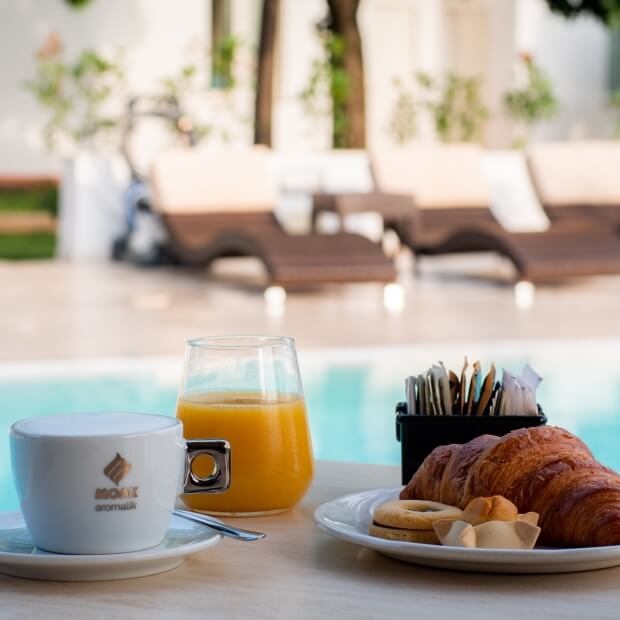 Tavolo con pasticcini, croissant, succo d'arancia e tazza, davanti a una piscina con sedie in vimini sullo sfondo.