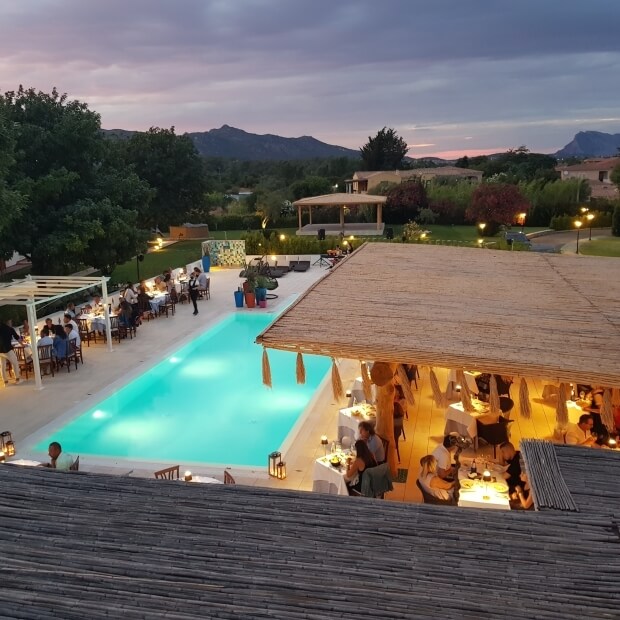 Immagine di un ambiente sereno con piscina turchese, ristorante, pergola bianca e case sullo sfondo, cielo nuvoloso al tramonto.