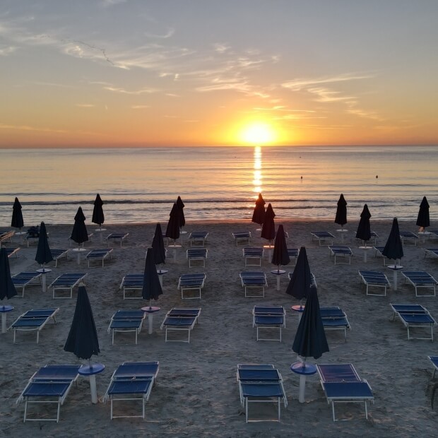 Spiaggia serena al tramonto con cielo arancione, mare calmo e sdraio e ombrelloni disposti in fila sulla sabbia.