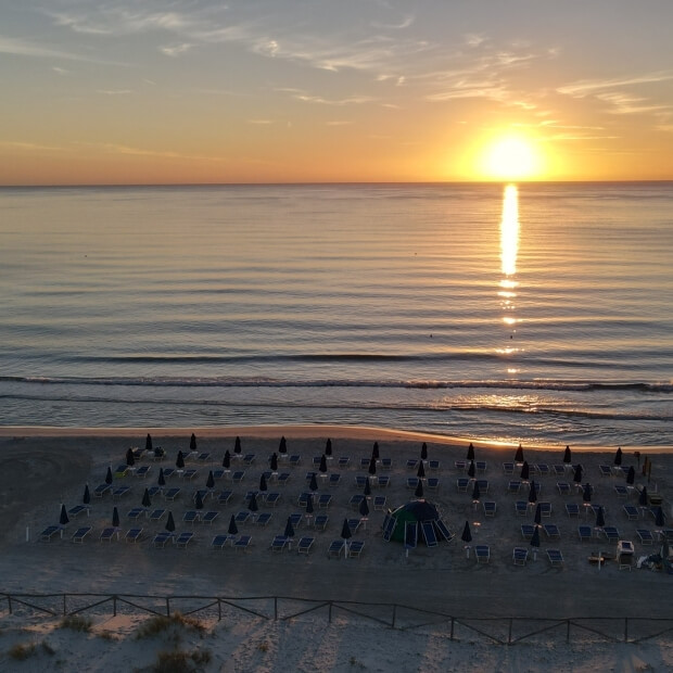 Spiaggia serena al tramonto con riflesso del sole sul mare, sedie a sdraio, ombrelloni e una recinzione nera.