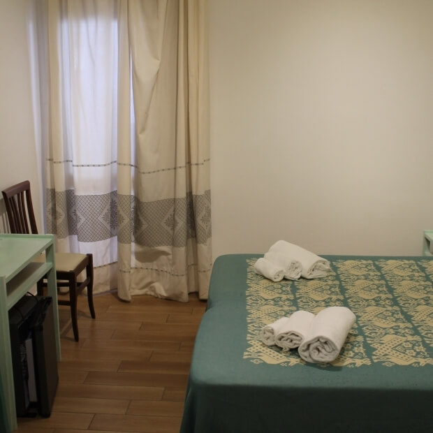 Camera d'hotel con letto, scrivania mint-verde e sedia, pareti bianche e pavimento in legno chiaro, atmosfera accogliente.