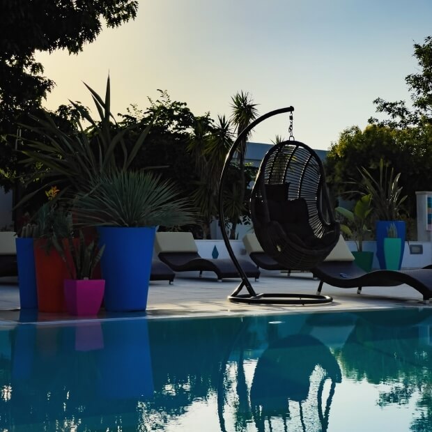 Una scena serena di piscina con un'amaca, sedie a sdraio, piante colorate e acqua turchese sotto un cielo blu e giallo.