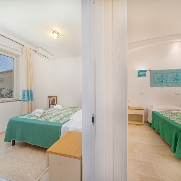 Interno di una stanza con due letti gemelli, pareti bianche, tappeto grigio e decorazioni semplici.