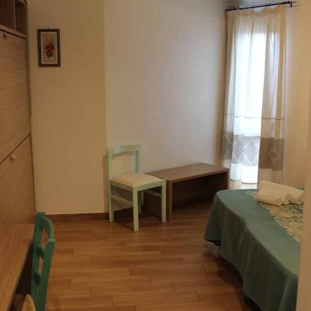 Una stanza minimalista e serena con pareti bianche, pavimento in legno, un tavolo, una sedia verde e un letto con lenzuola turchesi.