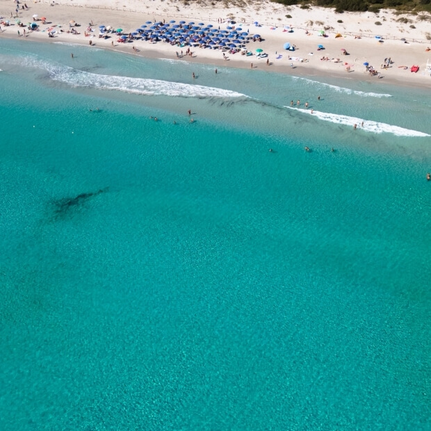 Una scena di spiaggia con persone che nuotano, acqua cristallina e ombrelloni, sotto un cielo soleggiato.