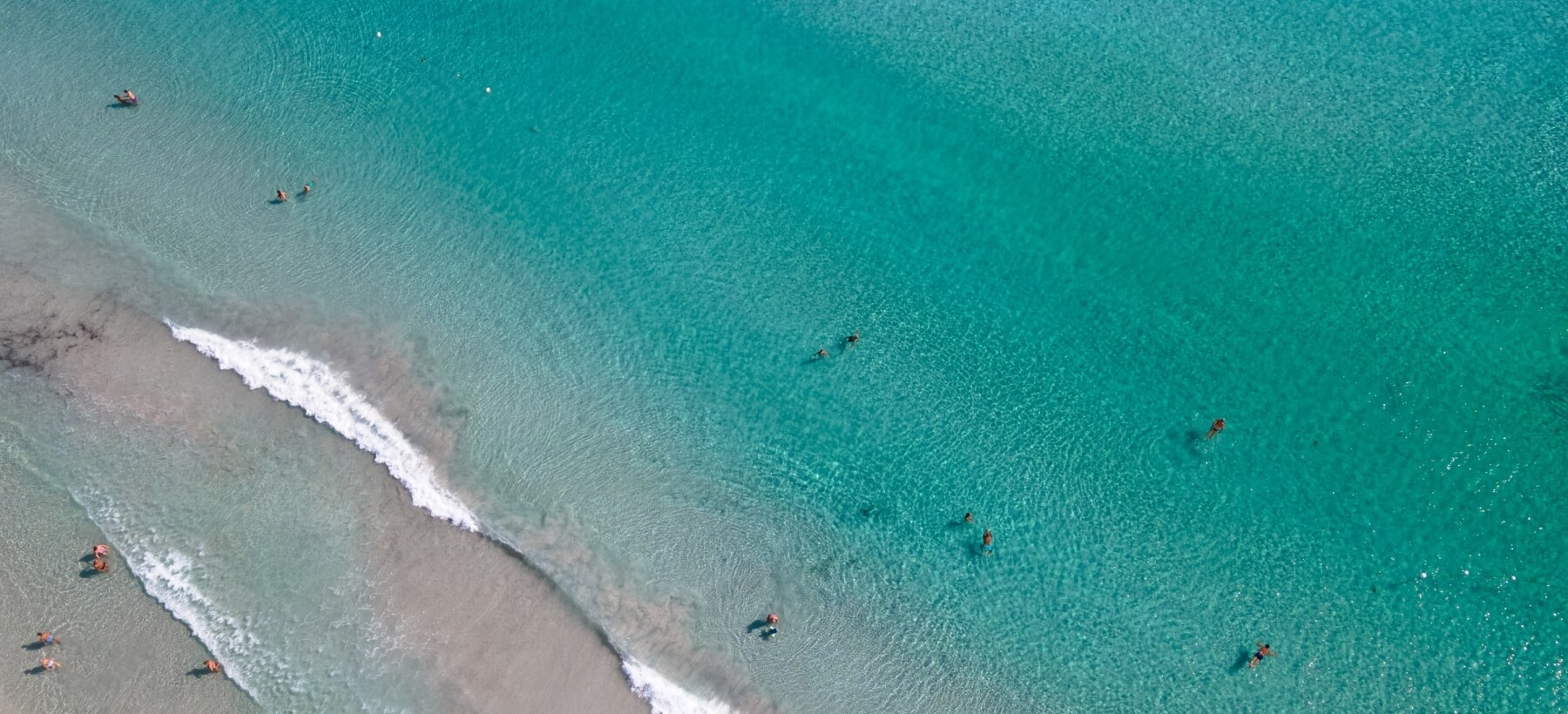 Spiaggia serena con acqua turchese, onde leggere e persone che nuotano, evocando tranquillità e relax estivo.