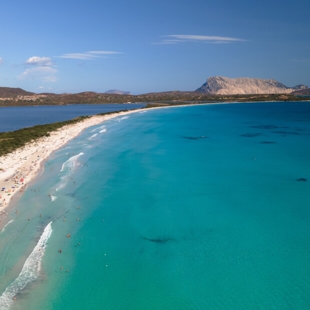 Una spiaggia serena con sabbia, persone che si godono il sole, onde gentili e una montagna sullo sfondo sotto un cielo blu.