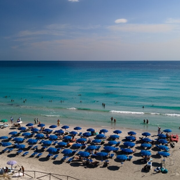 Una scena serena di spiaggia con ombrelloni blu, sabbia, oceano turchese e persone che si godono il sole e l'acqua.