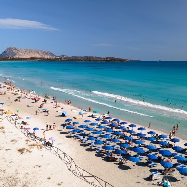 Spiaggia serena con ombrelloni blu, sabbia chiara, mare turchese e cielo blu punteggiato di nuvole. Atmosfera tranquilla.